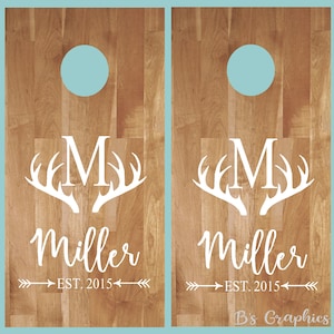 Op de afbeelding: Twee houten cornhole-borden met een wit monogram en een hertengewei-ontwerp. De borden hebben een lichtblauwe cirkel in het midden en de tekst "Miller EST. 2015" onder het ontwerp.