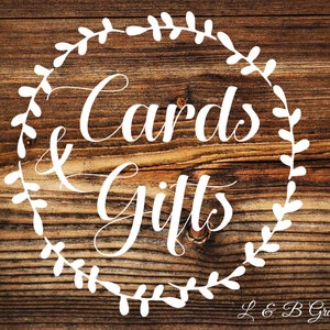Op de afbeelding: Een wit bord met een krans van bladeren rond de woorden "Cards & Gifts" op een bruine houten achtergrond. De tekst "L & B Graphics" staat in de rechterbenedenhoek.