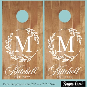 Op de afbeelding: Twee houten cornhole-borden met een lichtbruin houtnerfpatroon. Elk bord heeft een witte bloemenkrans rond een witte letter "M". De naam "Mitchell" en "EST. 2022" staan eronder. Bovenin een turquoise cirkel. Afmetingen: 50.8cm x 73.66cm.