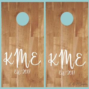 Op de afbeelding: Twee houten cornhole-borden met een lichtblauwe cirkel in het midden van elk bord. De borden hebben de initialen "KME" en de tekst "Est. 2017" in witte letters.