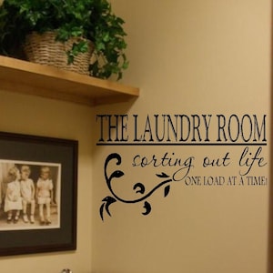 Op de afbeelding: Een zwarte en witte vinyl wand sticker met de tekst "The Laundry Room sorting out life one load at a time!" met een decoratief rankontwerp.