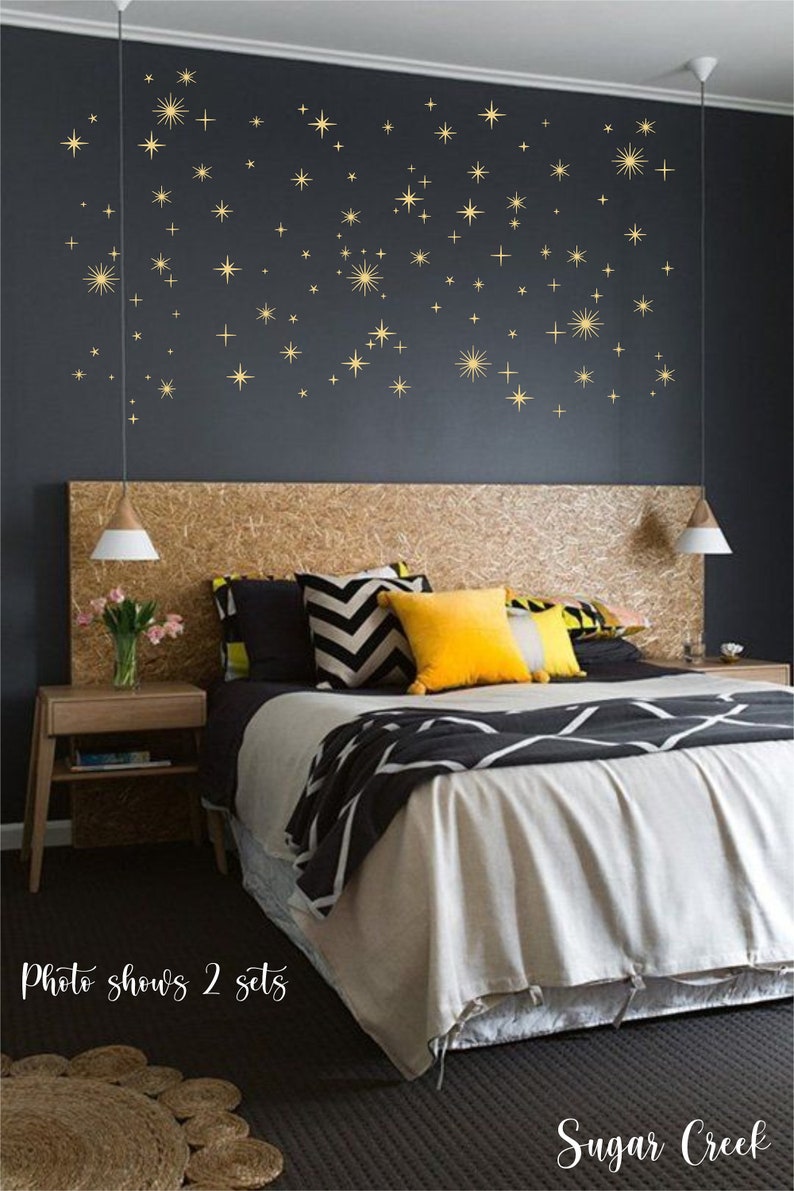 May include: A set of gold star wall decals featuring various sizes of four-pointed and multi-pointed star shapes arranged in a scattered pattern on a dark gray wall. The text "Photo shows 2 sets" and "Sugar Creek" is visible at the bottom.