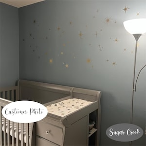 May include: A set of gold star-shaped wall decals applied to a blue-grey wall above a grey wooden crib and changing table combination. The decals feature various sizes of four-pointed and multi-pointed stars arranged in a scattered pattern.