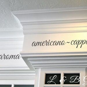 Op de afbeelding: Witte keukenkastjes met zwarte cursieve letters die "coffee - aroma" en "americano - cappuccino -" lezen. L & B Graphics staat in zwarte cursieve letters op de kastdeur.