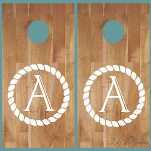 Op de afbeelding: Twee houten cornhole-borden met een wit touwontwerp en de letter "A" in het midden van elk bord. De borden zijn in een lichtbruine kleur geverfd en hebben een blauwe cirkel in het bovenste midden van elk bord.