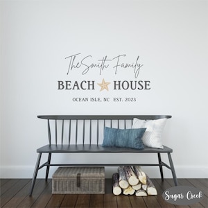 May include: A black and white wall decal with the text "The Smith Family Beach House" and a starfish graphic. The text "Ocean Isle, NC Est. 2023" is below the main text. The decal is displayed on a white wall above a black bench with two pillows.
