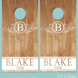 Op de afbeelding: Twee houten cornhole-borden met een wit monogramontwerp met de letter "B" en de naam "BLAKE" met het jaar "2016" eronder. De borden hebben een lichtbruine houtnerf.