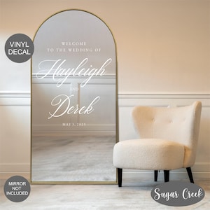 May include: A gold arched mirror with a white vinyl decal that says "Welcome to the wedding of Hayleigh and Derek May 3, 2025".
