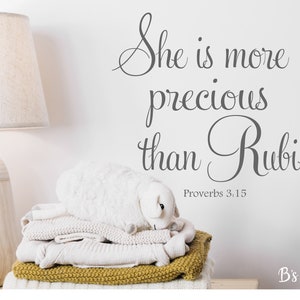 Può includere: Una lampada bianca con paralume beige è accanto a una pila di coperte piegate e un agnello di peluche. Il muro presenta il testo "She is more precious than Rubies" e "Proverbi 3:15".