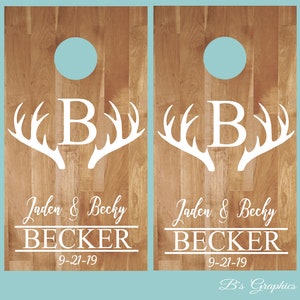 Op de afbeelding: Twee houten cornhole-borden met een wit hertengewei-ontwerp en de letter "B" in het midden. De borden hebben ook de namen "Jaden & Becky Becker" en de datum "9-21-19" erop gedrukt.