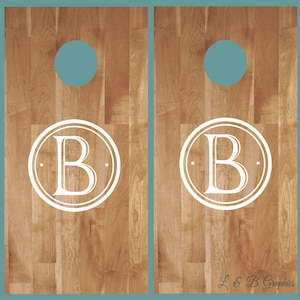 Op de afbeelding: Twee houten cornhole-borden met een lichtbruin houtnerfpatroon. Elk bord heeft een turquoisekleurig gat en een wit cirkelvormig ontwerp met de letter "B". De tekst "L & B Graphics" staat rechtsonder.