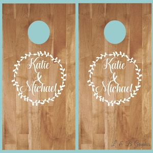Op de afbeelding: Twee houten cornhole-borden met een wit kransontwerp en de namen "Katie & Michael" in een cursief schrift.