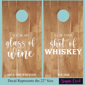 Op de afbeelding: Twee houten cornhole-borden met een lichtblauwe cirkel in het midden van elk bord. Het linker bord heeft de tekst "YOU BE MY glass of wine" en de tekst "MR & MRS WHEELER". Het rechter bord heeft de tekst "I'LL BE YOUR shot of WHISKEY" en de tekst "EST. 2020". De tekst "Decal Represents the 22" Size" staat onder de borden. De tekst "Sugar Creek" staat onder het rechter bord.