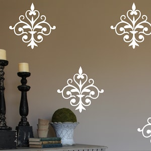 May include: White decorative wall decals in a classic, ornate design are displayed on a neutral-colored wall. The image also features black candle holders with lit candles, a topiary, and a decorative planter, creating a vintage aesthetic.