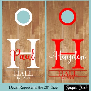 Op de afbeelding: Twee gepersonaliseerde cornhole-borden van 50,8 cm met witte en rode letters. Het linker bord zegt "Paul HALL Est. 2021" en het rechter bord zegt "Hayden HALL Est. 2021".