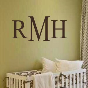 May include: A white crib with a brown and white patterned mattress and a white blanket. The wall behind the crib has a brown vinyl decal with the letters "RMH" on it.