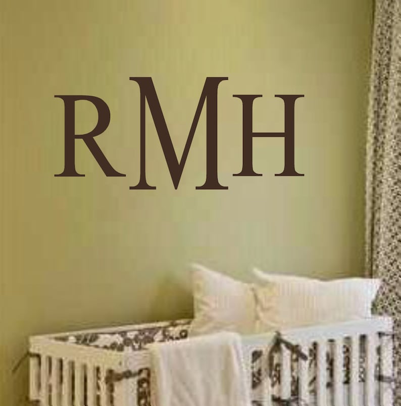 May include: A white crib with a brown and white patterned mattress and a white blanket. The wall behind the crib has a brown vinyl decal with the letters "RMH" on it.