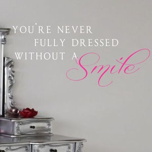 Puede incluir: Una pared blanca con un tocador plateado y una calcomanía de pared de vinilo rosa que dice "You're never fully dressed without a smile."