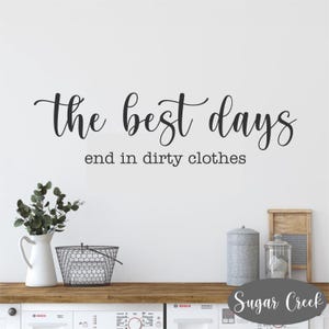 Könnte beinhalten: Eine weiße Wand mit schwarzem Text, der "the best days end in dirty clothes" lautet. Der Text ist in einer kursivschrift. Im Hintergrund befinden sich zwei Waschmaschinen.