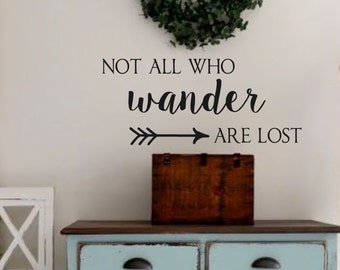 Puede incluir: Una calcomanía de pared en blanco y negro con la cita "Not all who wander are lost" y una flecha apuntando hacia la derecha. La calcomanía está en una pared blanca sobre una cómoda azul claro con dos cajones.