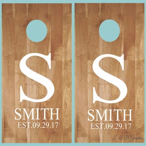 Op de afbeelding: Twee houten cornhole-borden met een lichtbruin houtnerfpatroon. Elk bord heeft een grote witte letter "S", de naam "SMITH" en de datum "EST.09.29.17". Een lichtblauwe cirkel staat bovenaan elk bord.