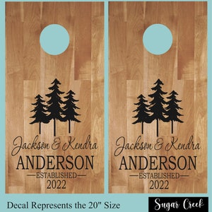 Könnte beinhalten: Zwei rechteckige Cornhole-Bretter aus Holz mit einer hellbraunen Holzmaserung und einem kreisförmigen Loch an der Oberseite. Jedes Brett zeigt eine schwarze Grafik von drei Kiefern über dem Text Jackson & Kendra ANDERSON ESTABLISHED 2022.