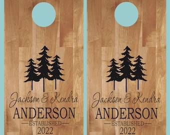 Könnte beinhalten: Zwei rechteckige Cornhole-Bretter aus Holz mit einer hellbraunen Holzmaserung und einem kreisförmigen Loch an der Oberseite. Jedes Brett zeigt eine schwarze Grafik von drei Kiefern über dem Text Jackson & Kendra ANDERSON ESTABLISHED 2022.