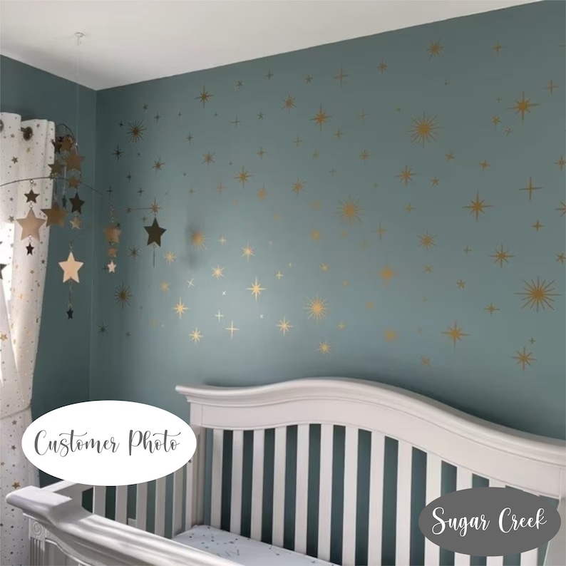 May include: A set of gold star wall decals applied to a muted blue wall above a white crib. The decals feature various sizes of four-pointed and multi-pointed stars, creating a scattered celestial pattern across the flat surface of the wall.