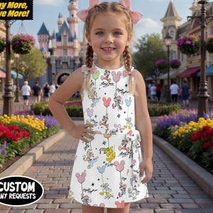May include: A white tennis dress skirt with a Disney theme, featuring Mickey Mouse and friends with balloons. The dress has a wrap-around tie and is worn by a young girl. The background shows a theme park setting with flowers and buildings.