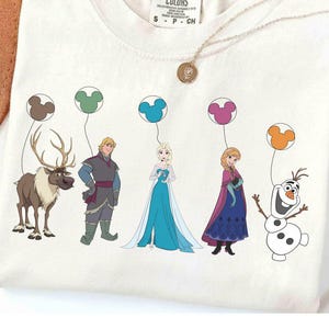 May include: Cream-coloured Comfort Colors t-shirt featuring a colourful graphic of characters from the animated film Frozen. The characters are holding Mickey Mouse-shaped balloons. A gold necklace and a brown hat are also visible.