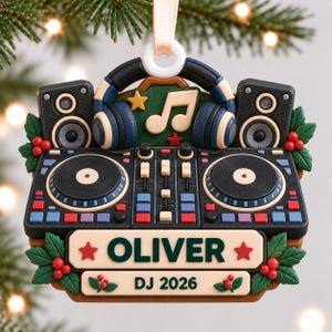 Peut inclure: Une décoration de Noël représentant une console de DJ noire, deux haut-parleurs et un casque bleu. Une note de musique et une étoile sont centrées au-dessus. Une plaque crème affiche le texte OLIVER et DJ 2026, ornée de houx vert et de baies rouges.