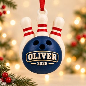 May include: A bowling-themed Christmas ornament featuring three white bowling pins with red stripes positioned behind a dark blue bowling ball. The ball displays the name OLIVER and the year 2026 in raised white lettering on a gold-bordered plaque.