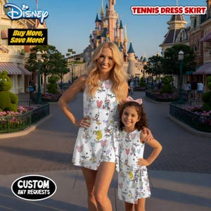 May include: A woman and a child wearing matching white tennis dress skirts with a Disney character balloon print. The dresses have a wrap-style skirt and are sleeveless. The background features a Disney theme park with a castle.