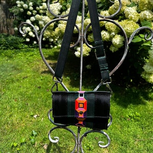 May include: A small black crossbody bag with a green strap and colorful geometric design. The bag is hanging on a metal stand.