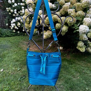 May include: A teal blue bucket bag with a drawstring closure and a long strap. The bag is made of a striped fabric and has a silver metal buckle on the strap.
