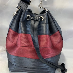 Puede incluir: Un bolso de cubo a rayas negras y burdeos con una correa ajustable negra y un cierre de cordón.