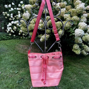 Puede incluir: Un bolso de cubo rosa con un cordón de ajuste rojo y una correa roja. El bolso está hecho de un material tejido y tiene un efecto degradado rosa claro.