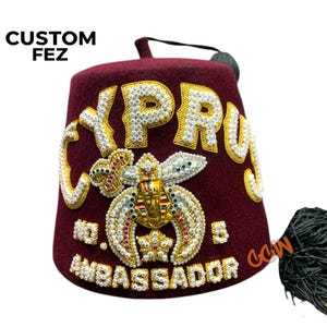 Puede incluir: Un fez granate con bordados dorados y pedrería. El fez tiene la palabra "CYPRUS" bordada en la parte delantera, junto con un símbolo masónico y el texto "AMBASSADOR".