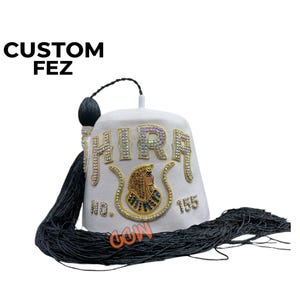 May include: A white fez hat with the word "HIRA" in jeweled letters. The hat features a gold and jeweled emblem and a black tassel. The text "CUSTOM FEZ" is at the top left.