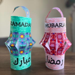 May include: Two paper lanterns decorated with colorful patterns and the words "Ramadan Mubarak". The lantern on the left is green with purple and yellow accents, and the lantern on the right is pink with red and white accents.