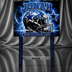 May include: A rectangular sign featuring a blue and white football helmet graphic with stars, the text JONAH at the top, and COWBOYS at the bottom. The sign is supported by a blue metal stand with a horizontal bar displaying the name Jonah in black text.