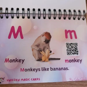 Può includere: Una scheda educativa con rilegatura a spirale che mostra la lettera M in maiuscolo e minuscolo, una foto di una scimmia che sbuccia una banana, il testo Monkey, monkey e la frase Monkeys like bananas. Sono presenti un codice QR e Unicorn Magic Cards.