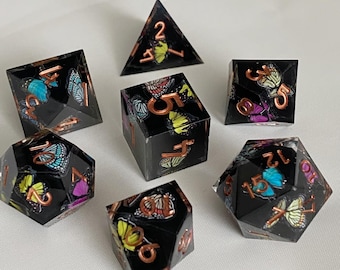 May include: Set of seven black dice with colorful butterfly designs and copper-colored numbers. The dice are a d4, d6, d8, d10, d%, d12, and d20.