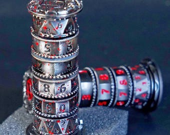 May include: Two silver-coloured, cylindrical dice towers with red numbering and accents. The towers are stacked with rotating rings displaying numbers. The top and bottom have decorative detailing. The dice towers are on a dark surface.