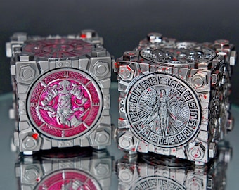 May include: Two metallic, cube-shaped puzzle boxes with detailed designs. One has a pink circular emblem with a figure, the other a silver emblem with an angel-like figure. Both have a silver finish with red accents.