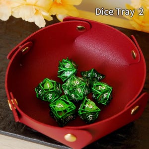May include: A red leather dice tray with gold rivets contains several green metal dice. The dice feature a leaf design and white numbers. The text "Dice Tray 2" is visible in the upper right corner.
