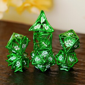 May include: Three green metal dice with white numbering and leaf designs. The dice are in various geometric shapes, including a pyramid and two multi-sided dice. The dice are arranged on a dark surface.