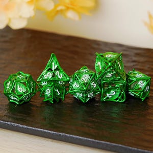 May include: A collection of six green metal dice featuring a leaf design. The dice are in different geometric forms, such as a d4, d6, d8, d10, d12, and d20. The numbers are white and clearly visible.