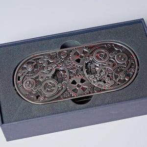 May include: A silver-colored belt buckle with a complex gear design and red accents. The buckle is rectangular with rounded ends and is presented in a dark gray box. The gears are intricately detailed, creating a steampunk aesthetic.