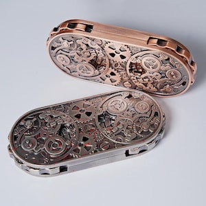 May include: Two steampunk-style metal cases with intricate gear designs. One case is bronze-colored, the other is silver with red accents. The cases are rectangular with rounded ends, showcasing a complex mechanical aesthetic.